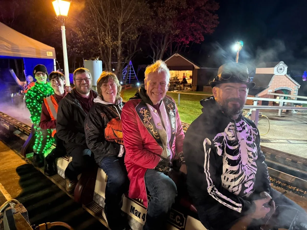 Passengers, several in Halloween costume including a red ringmaster's coat, a skeleton top and a light-up suit, sitting astride a miniature train at night
