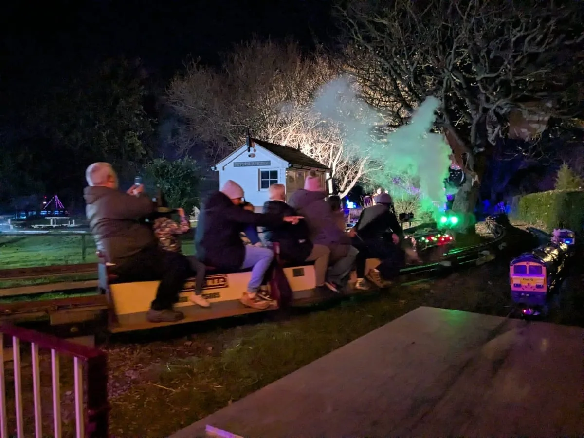 A miniature train full of passengers passing a lit station building at night, with steam rising among the trees