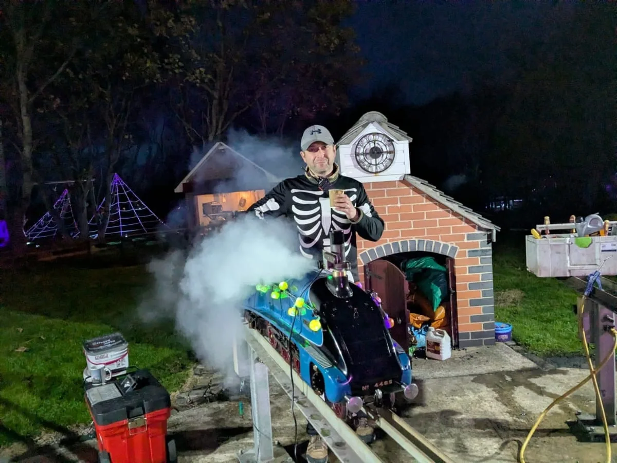 A driver in a skeleton costume and cap standing behind a blue miniature steam locomotive decorated with coloured lights, steam around it, beside a brick station with a clock