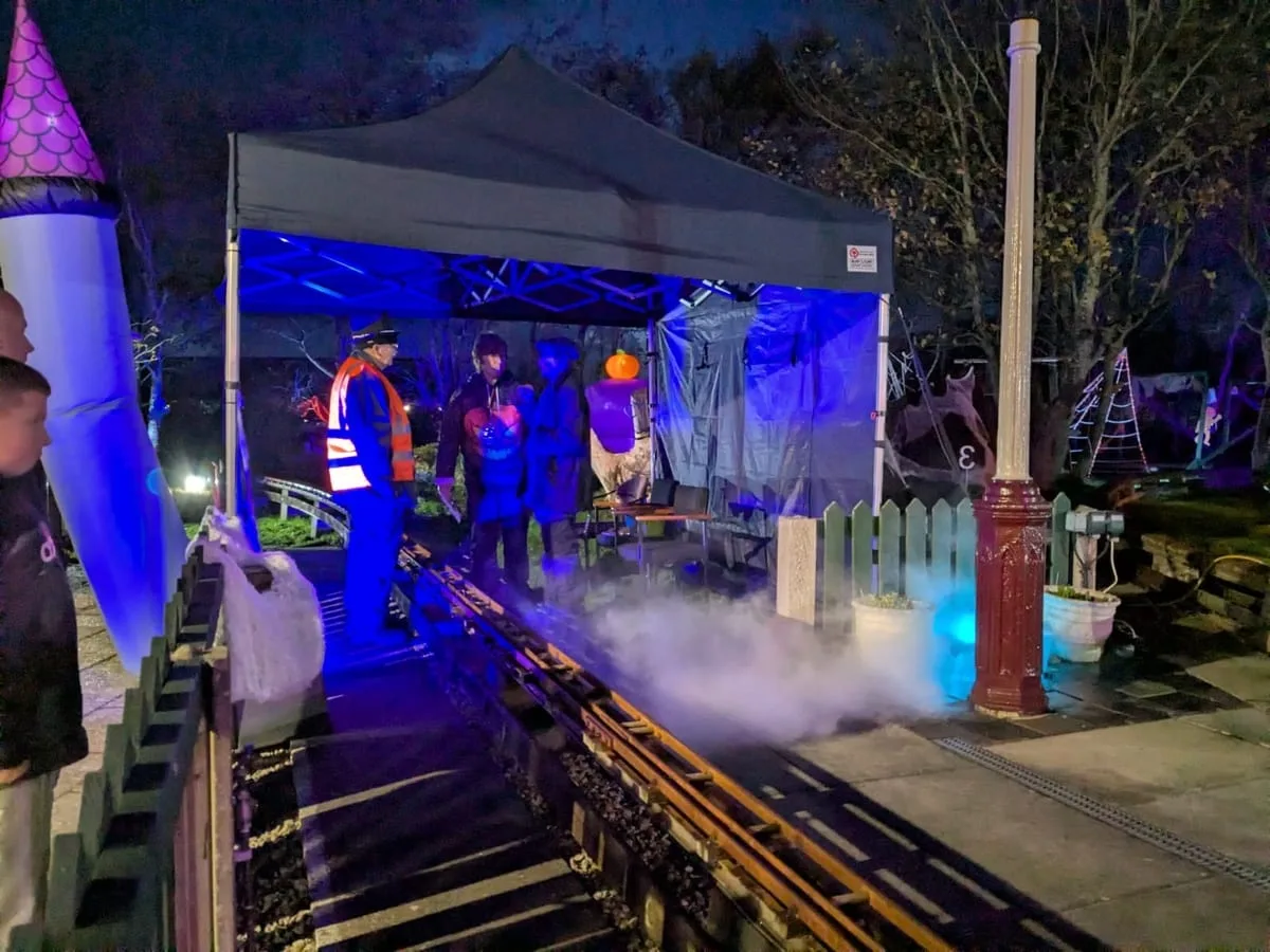 A miniature railway platform at night under blue light, with low fog across the track, a steward in hi-vis and people in costume under a gazebo