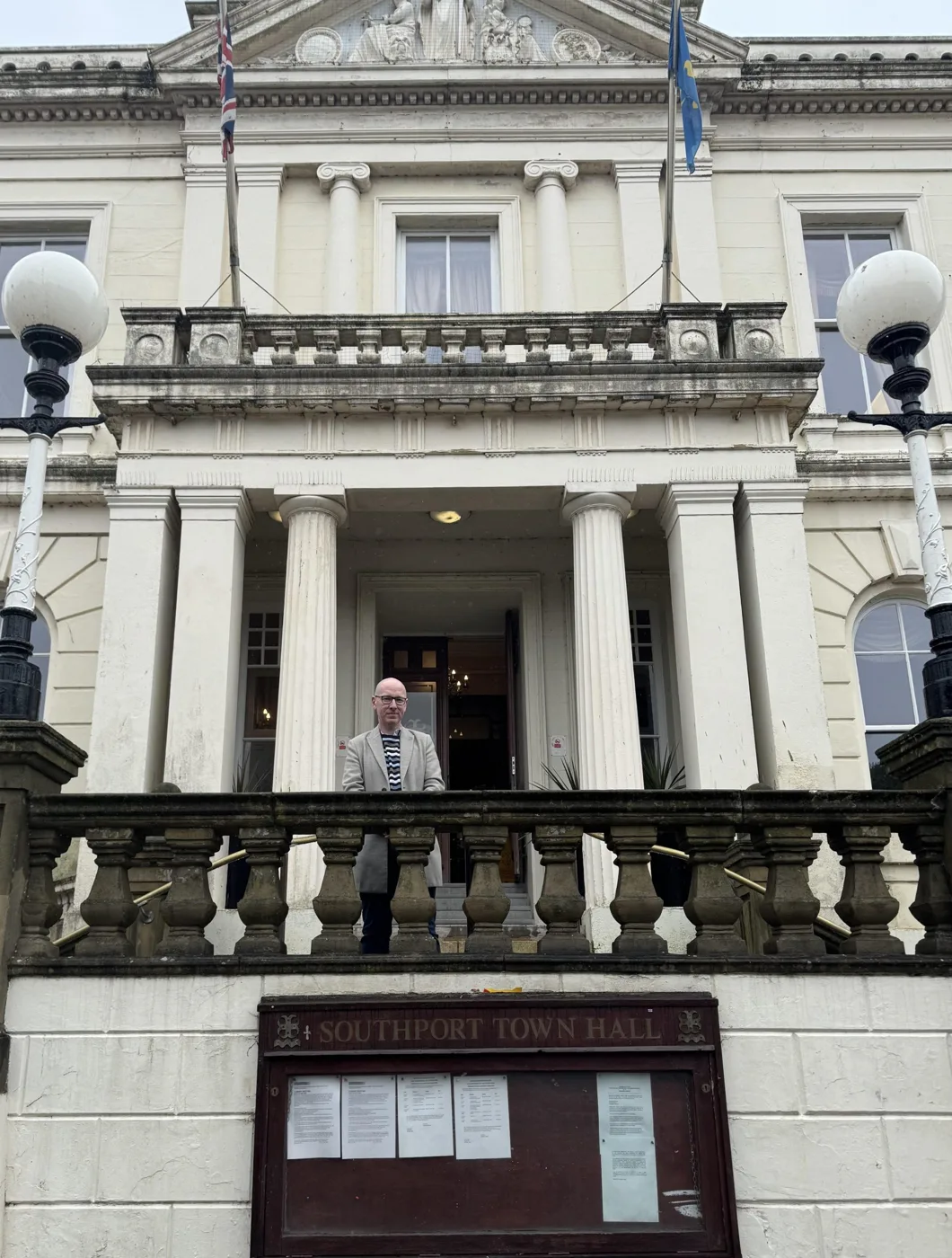 Patrick Hurley standing on the steps of Southport Town Hall behind the balustrade, above the Southport Town Hall notice board
